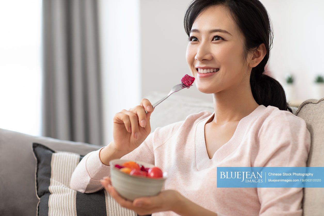 Happy young Chinese woman eating fruit salad in living room