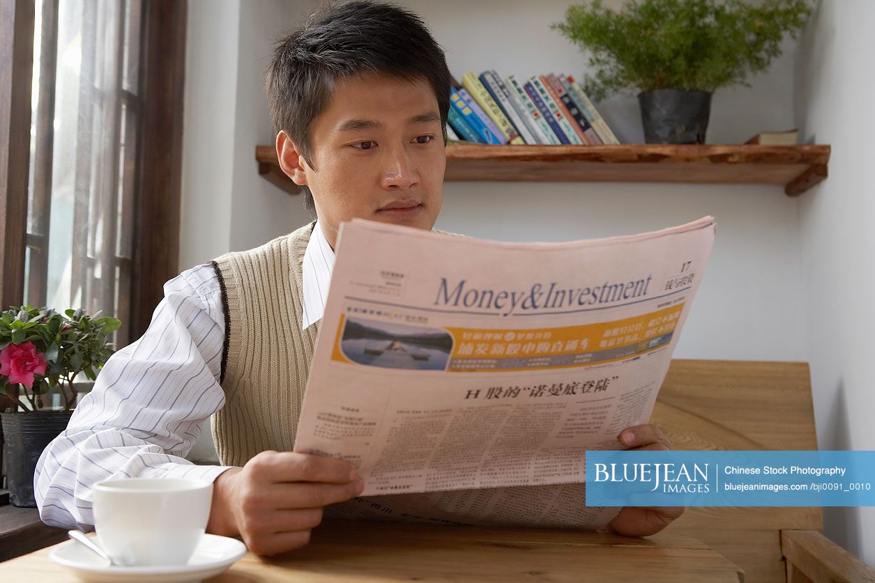 Chinese Man Sitting At Table, Reading A Newspaper