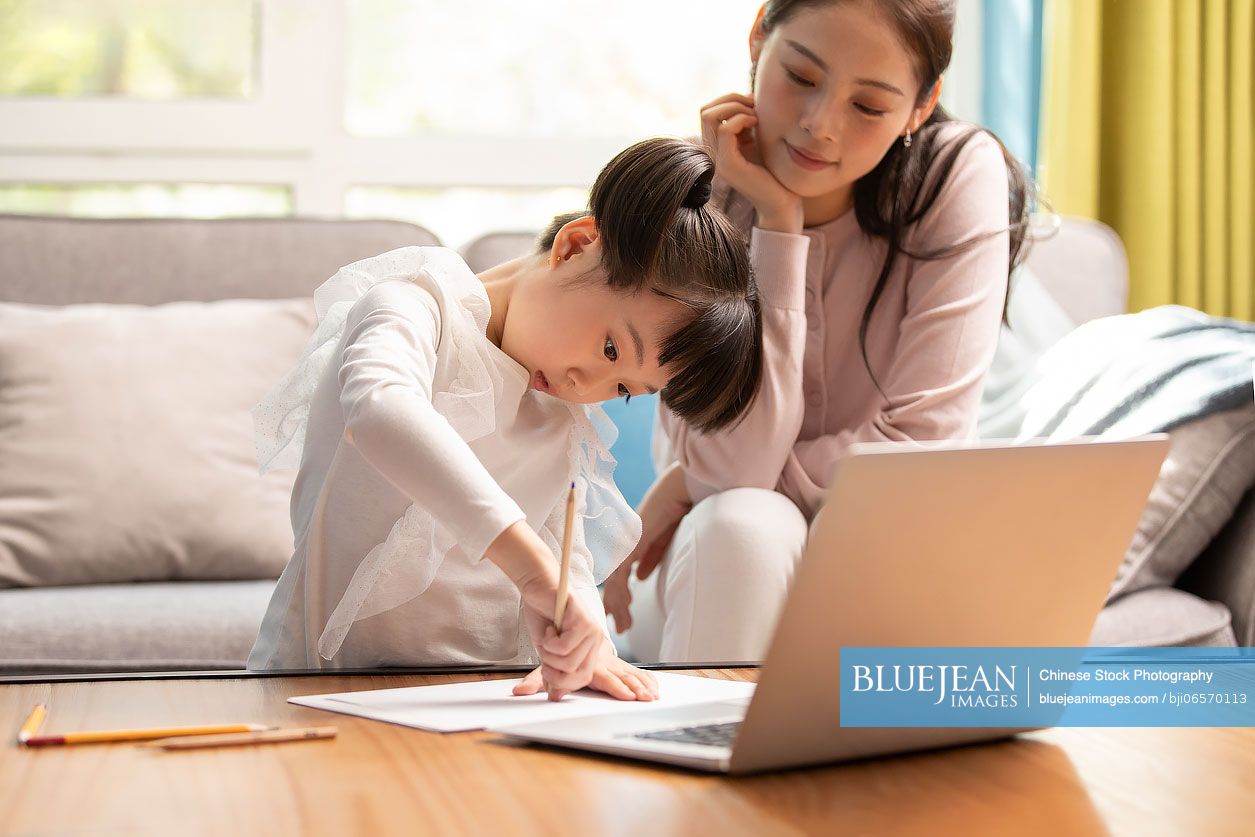 Young Chinese mother helping daughter with her homework in living room