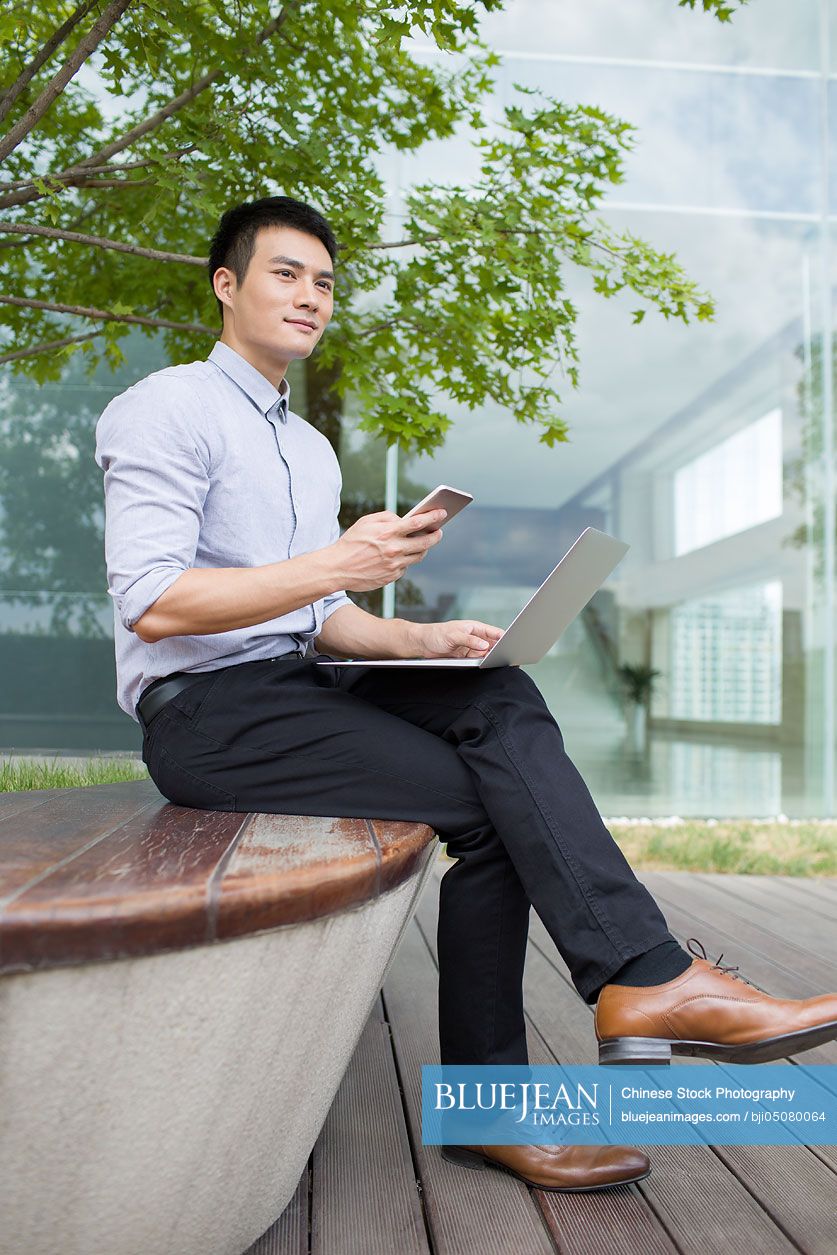 Young Chinese businessman using laptop and smart phone outdoors