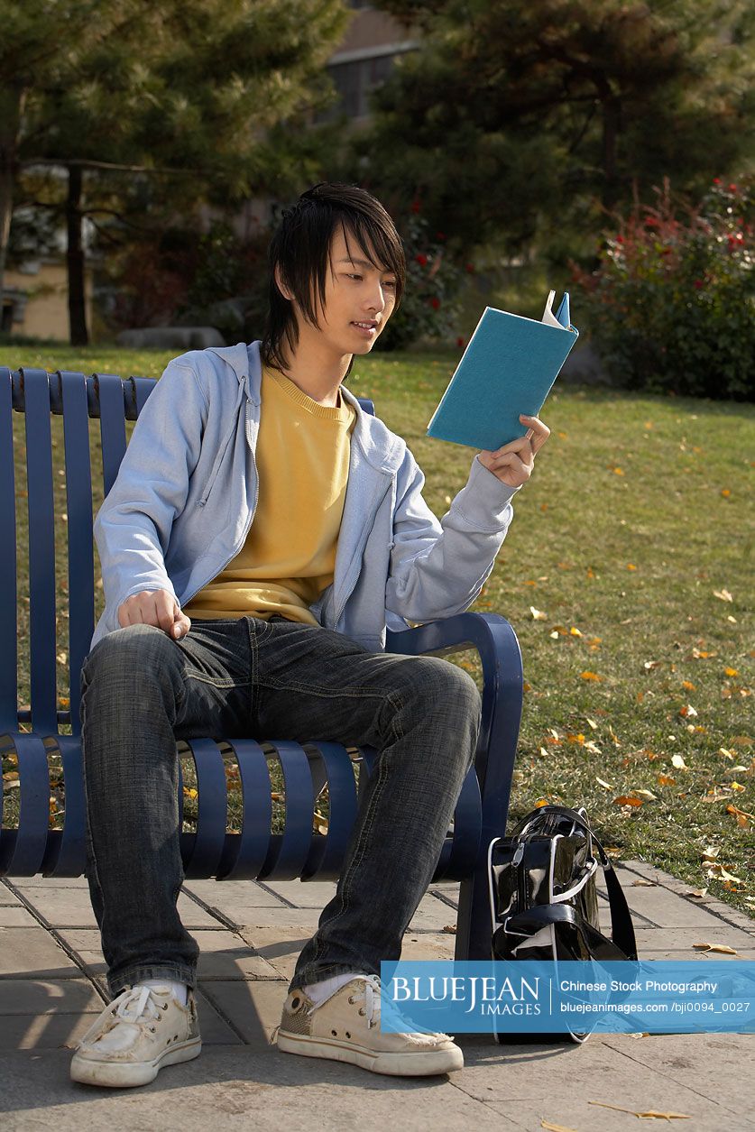 Young Chinese man sitting on park bench reading book