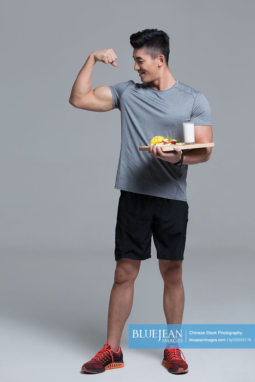 Young Chinese male athlete flexing muscles while holding vegetable salad and milk