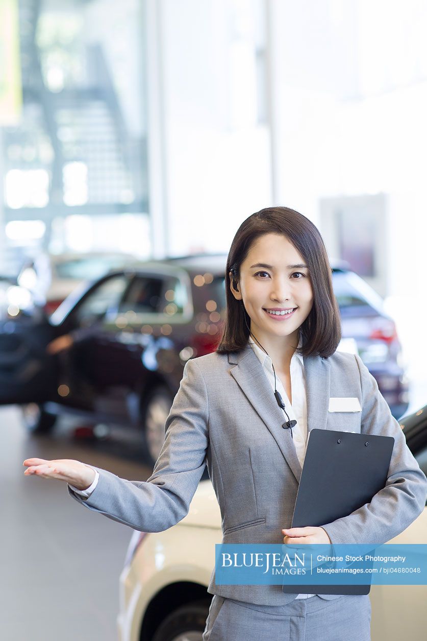 Confident Chinese saleswoman standing with new cars in showroom