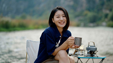 Cheerful young Chinese woman enjoying hot drink while sitting on camping chair