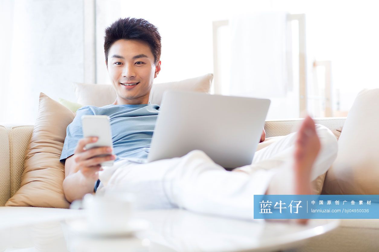 年轻男子在沙发上使用笔记本电脑和手机