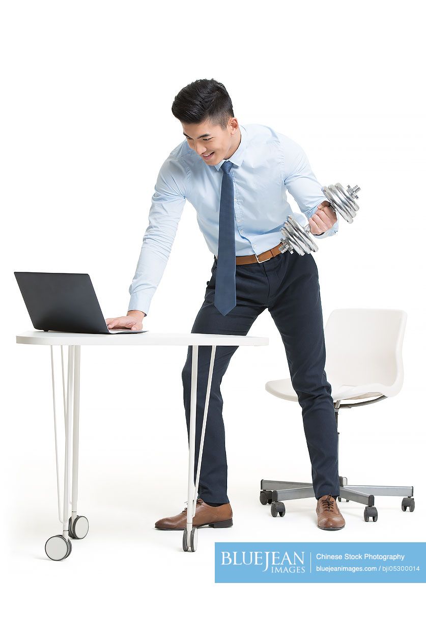 Young Chinese businessman lifting weights while using laptop at work