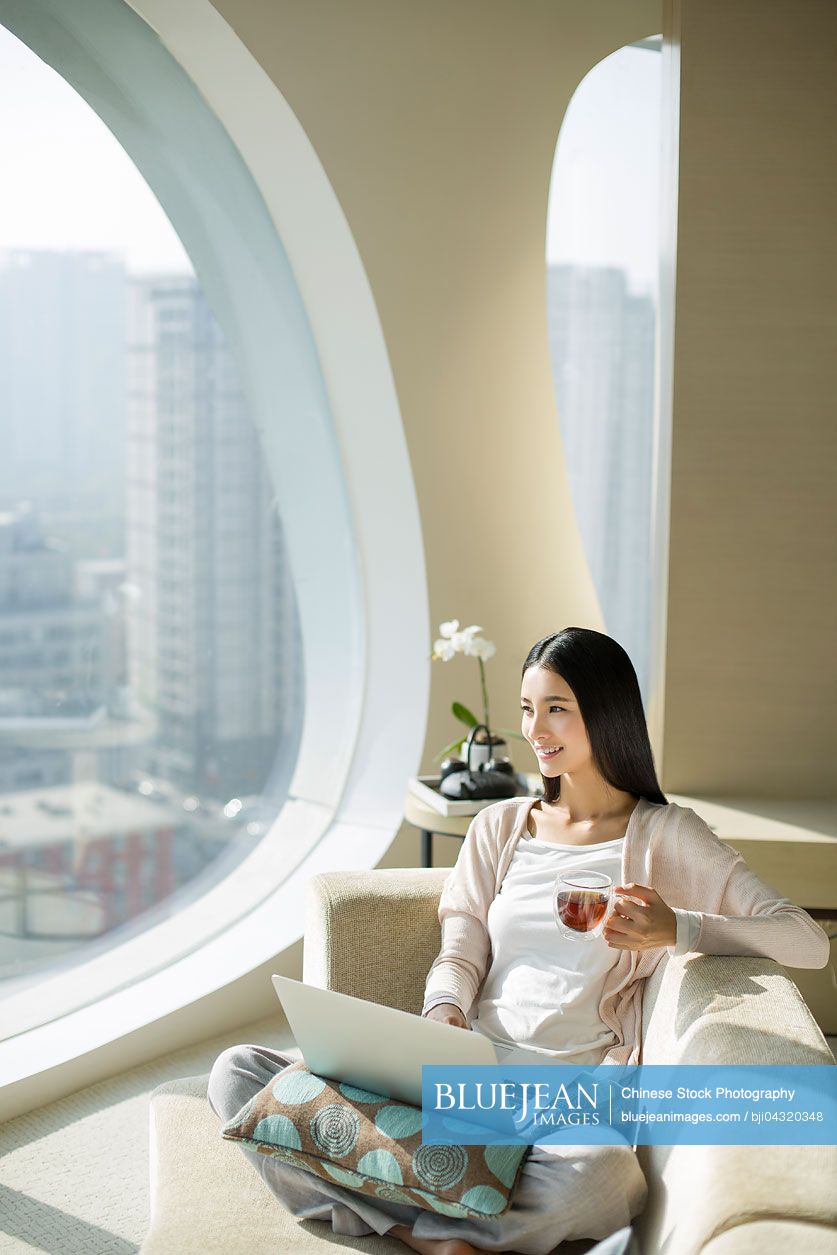 Young Chinese woman with tea cup using laptop on couch