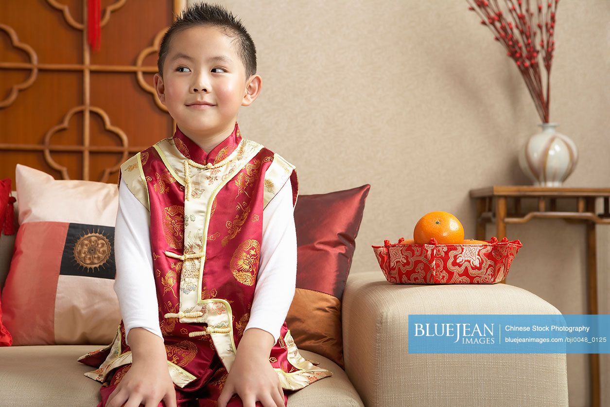 Young Chinese boy sitting on couch in traditional clothing looking mischievous