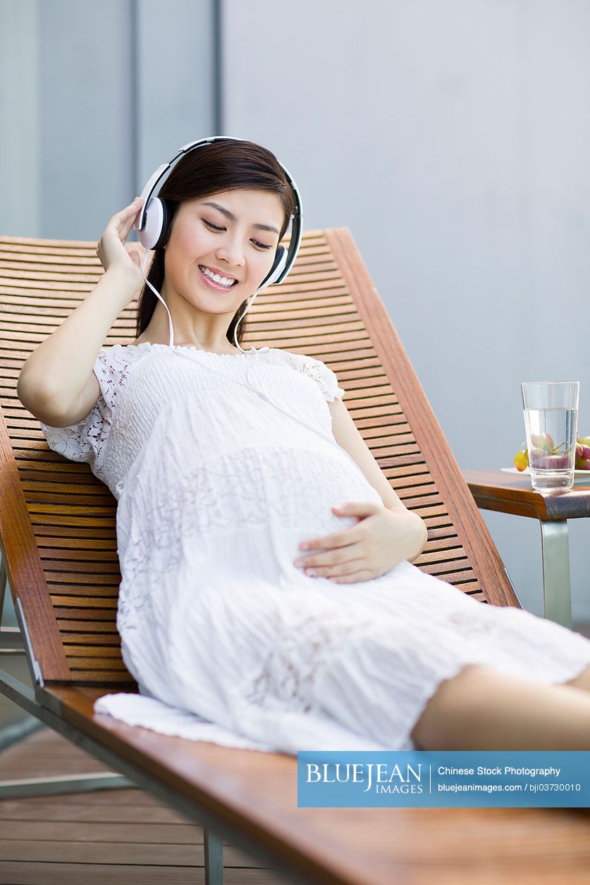 Pregnant Chinese woman listening to music on chaise longue