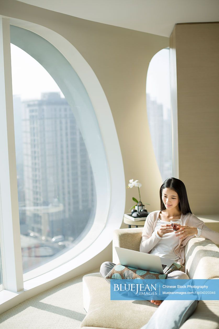 Young Chinese woman with tea cup using laptop on couch