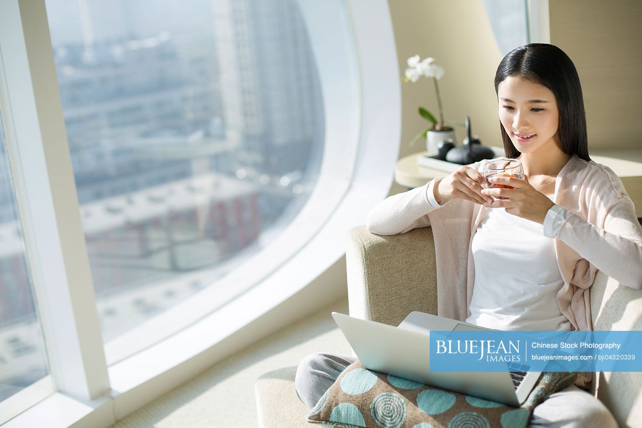 Young chinese woman with tea cup using laptop on couch high res stock