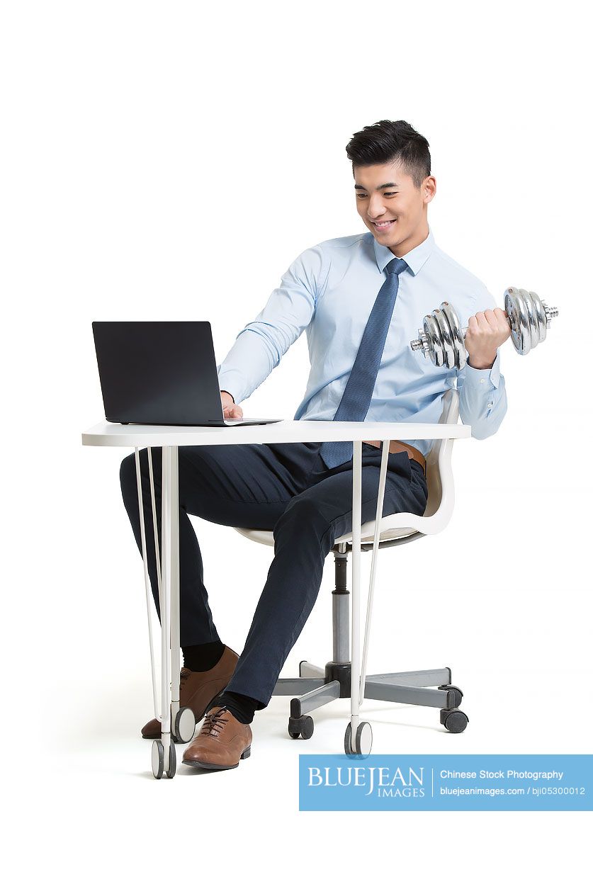 Young Chinese businessman lifting weights while using laptop at work