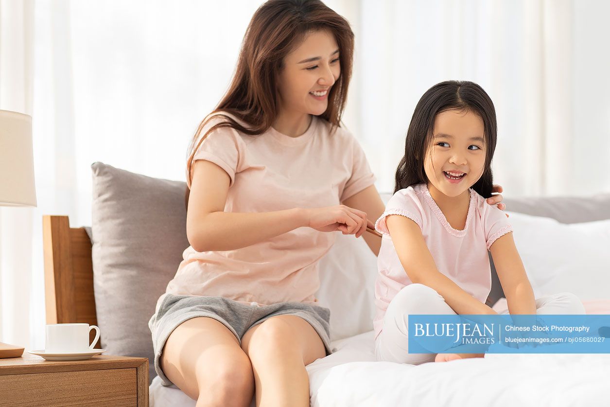 Happy Chinese mother combing her daughter's hair in bedroom