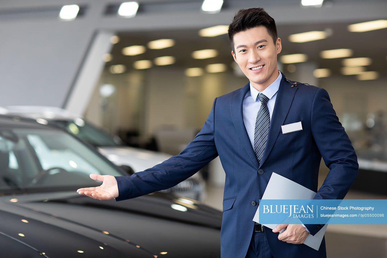 Confident Chinese salesman with cars in showroom