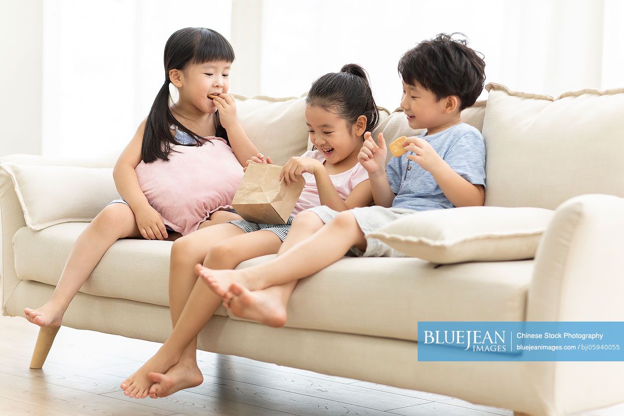 Cute Chinese children eating biscuits on sofa