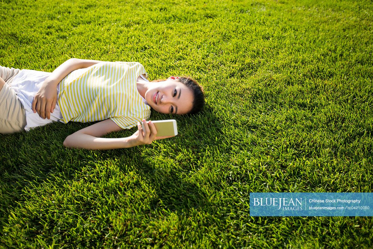 Cheerful young Chinese woman lying on grass with cell phone