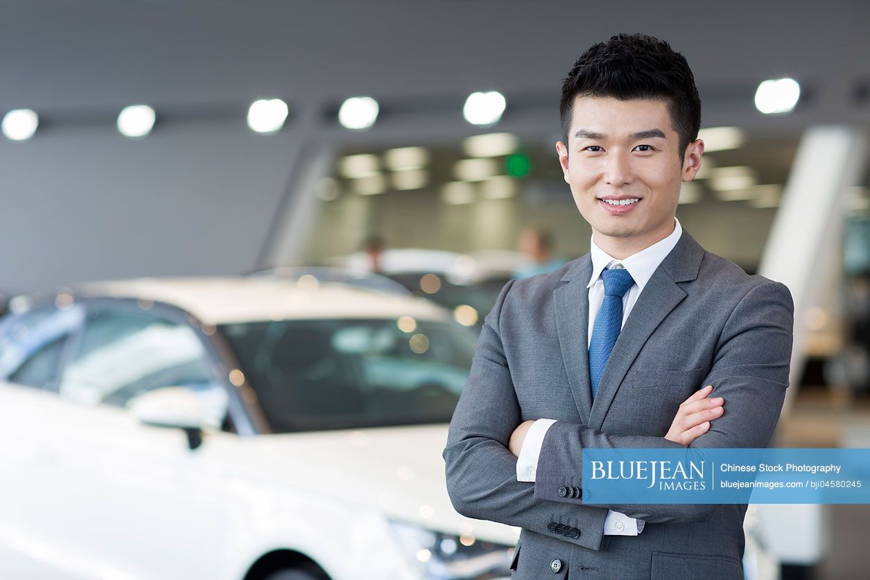 Confident Chinese salesman standing with new cars in showroom
