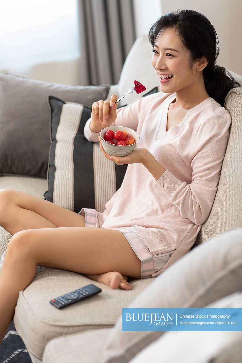 Happy young Chinese woman eating fruit salad in living room