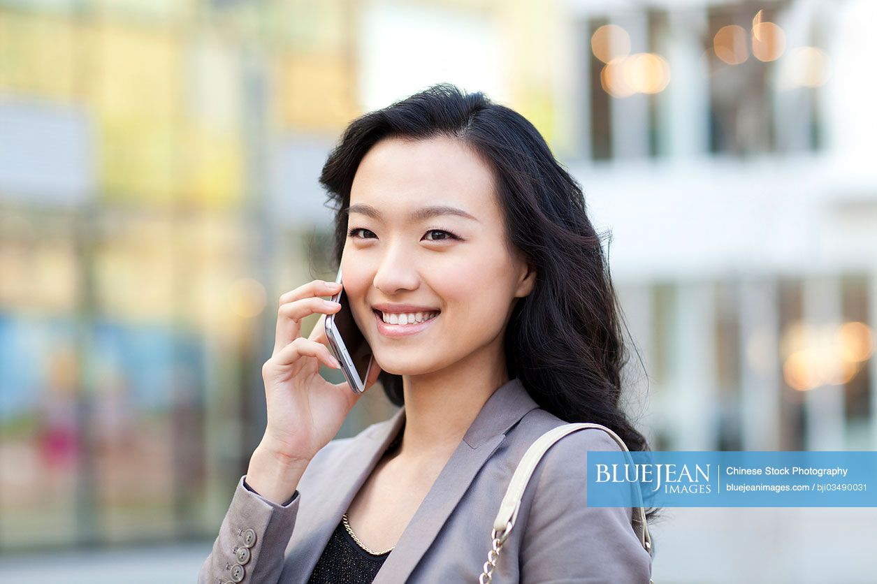 Cheerful young Chinese woman talking on the phone in city