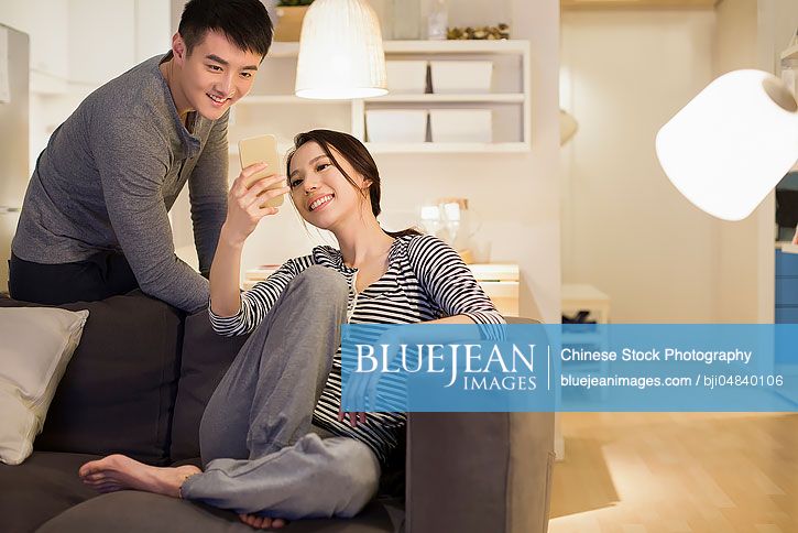 Young Chinese couple using smart phone on living room sofa