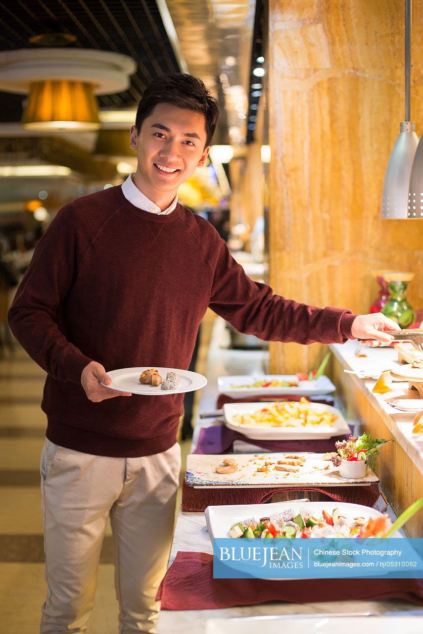 Cheerful young Chinese man taking food from buffet table