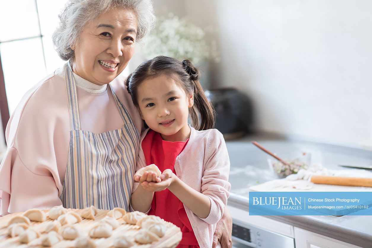 Cheerful Chinese granddaughter and grandmother making dumplings in kitchen