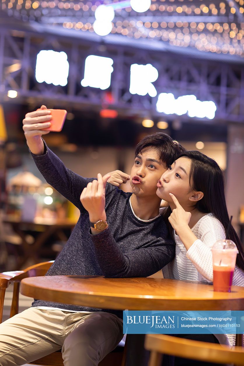 Happy young Chinese couple taking selfie with smartphone in shopping mall