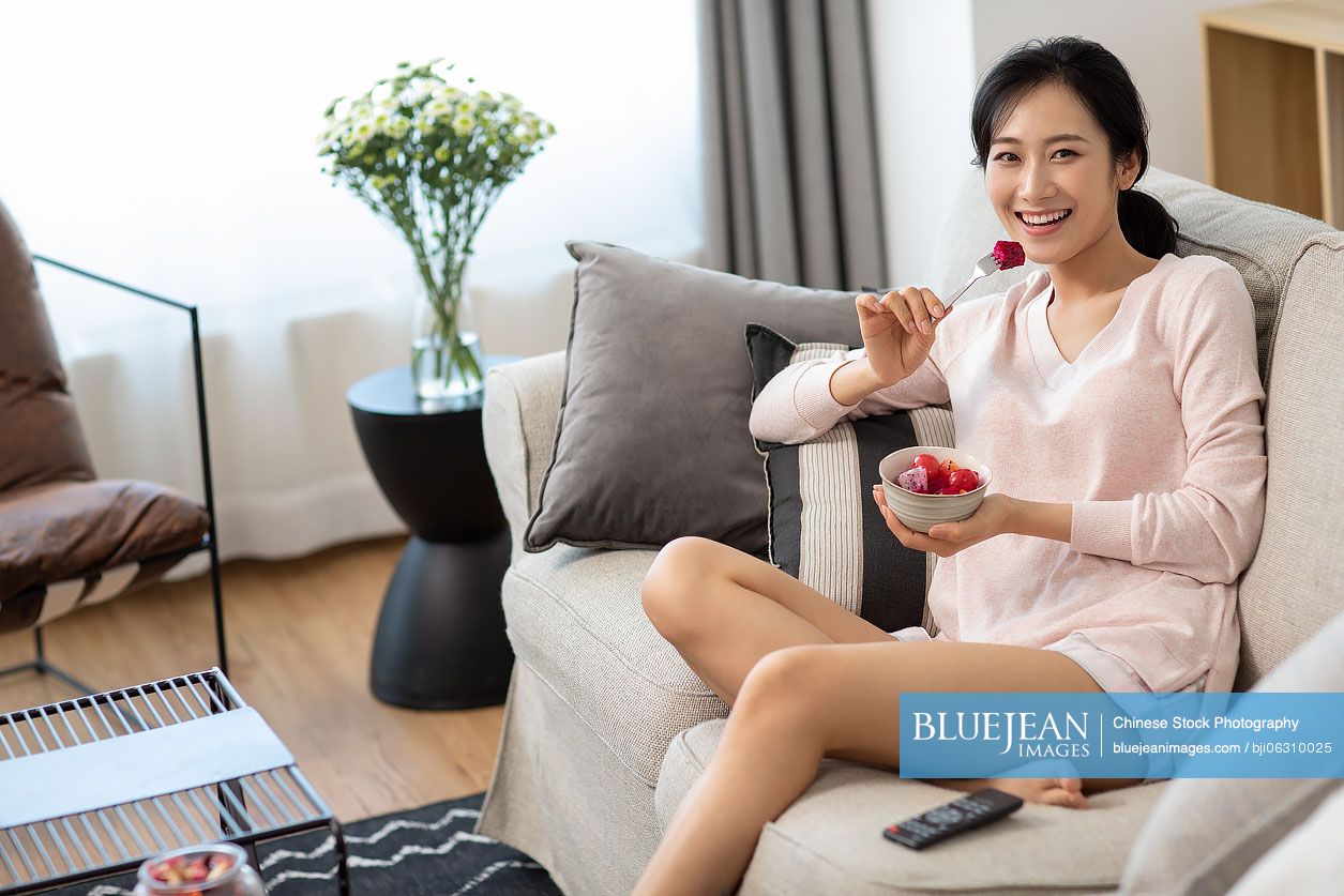 Happy young Chinese woman eating fruit salad in living room