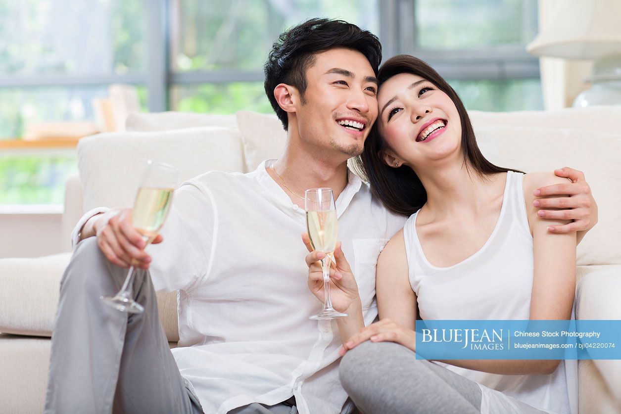 Cheerful young Chinese couple drinking champagne in living room