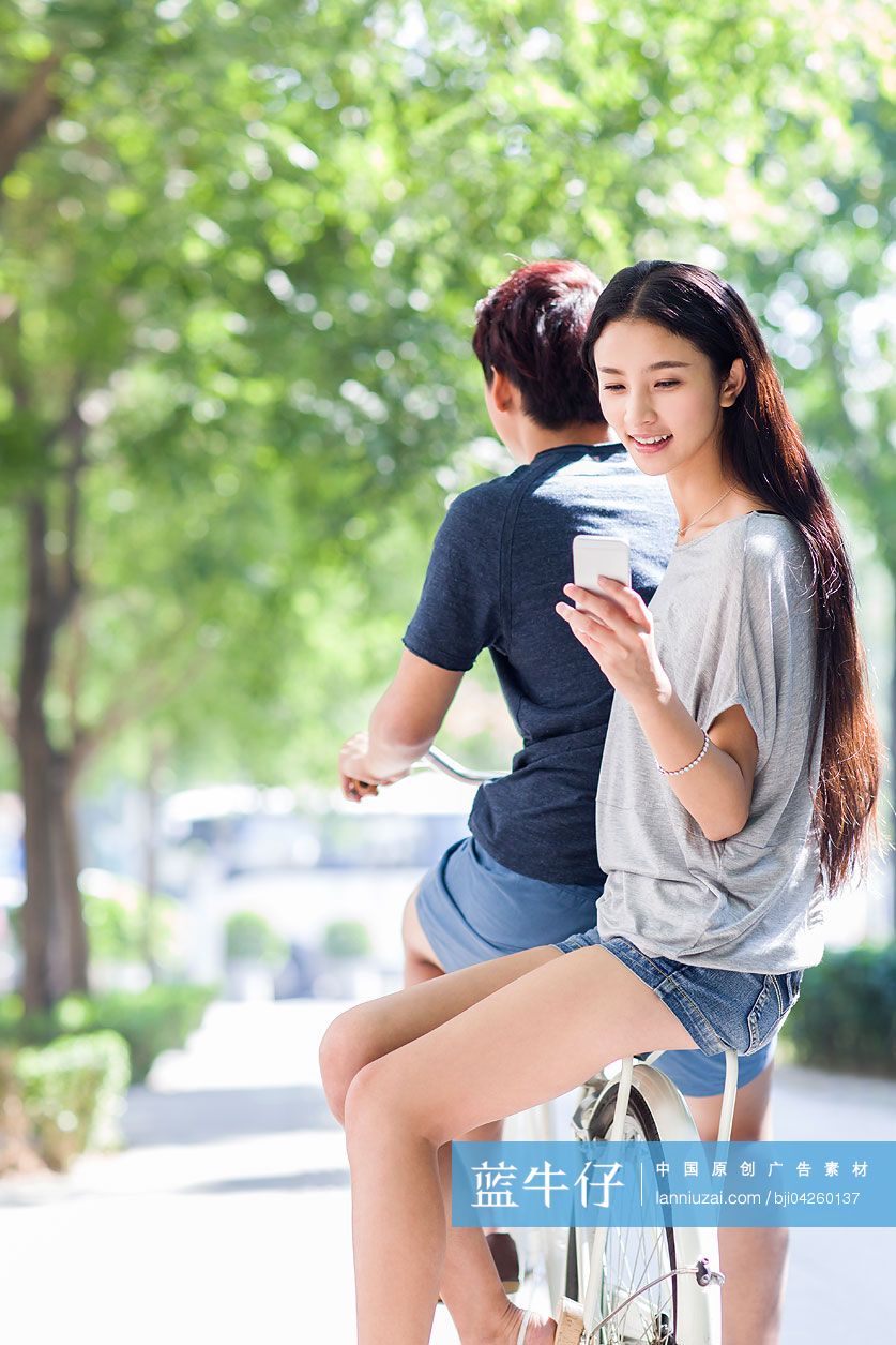 年轻女子坐在男友自行车后面玩手机