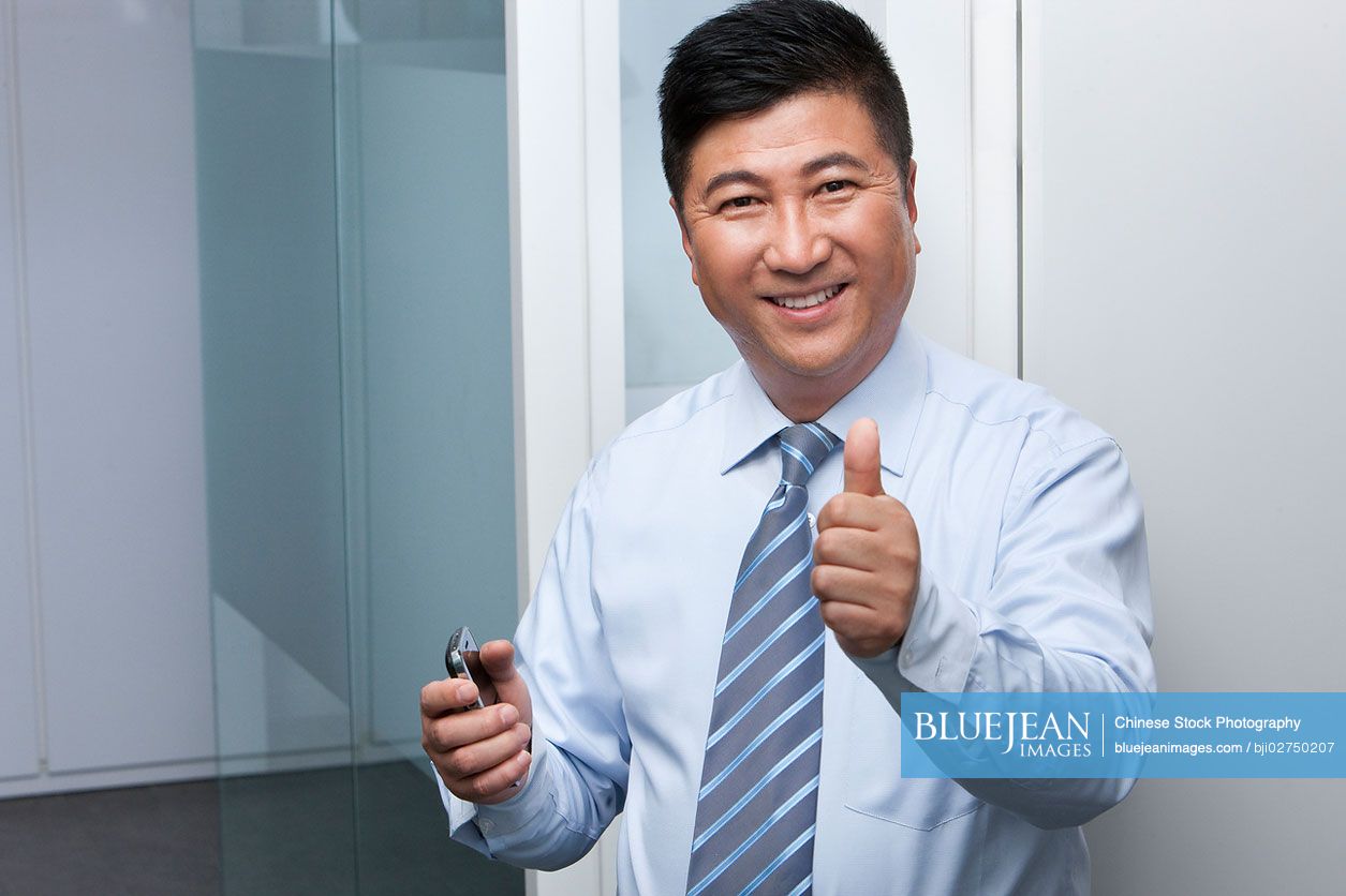 Smiling Chinese businessman holding his mobile phone giving the thumbs-up signal