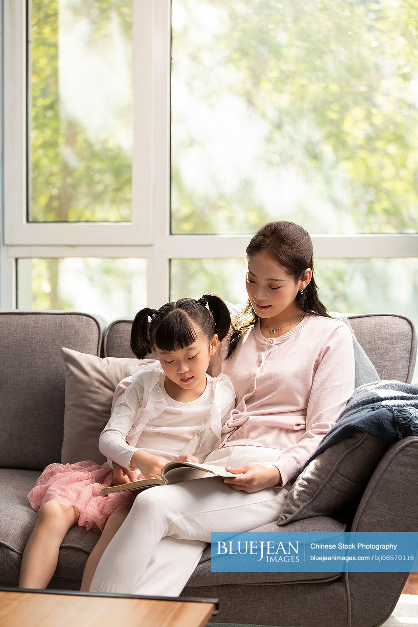 Young Chinese mother and daughter reading book at home