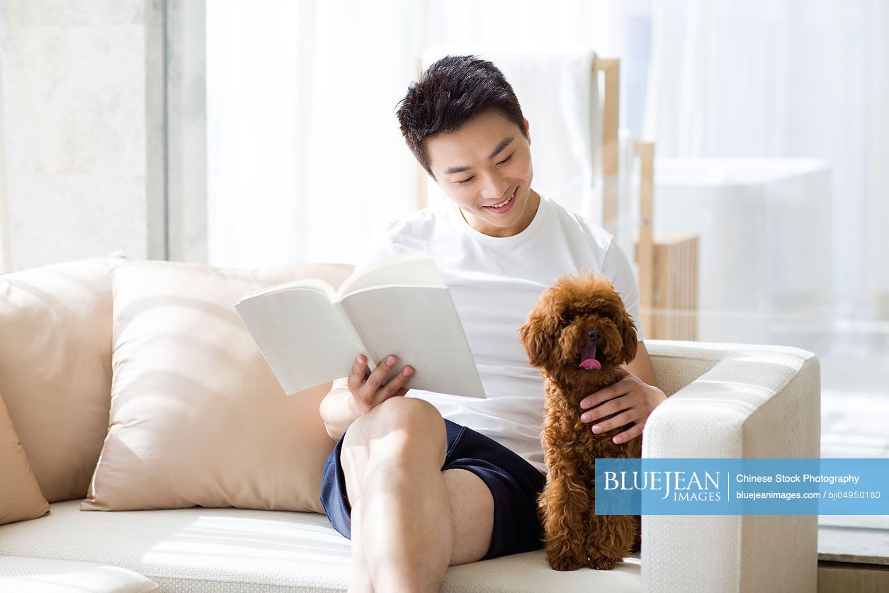 Young Chinese man reading with a pet poodle on the sofa