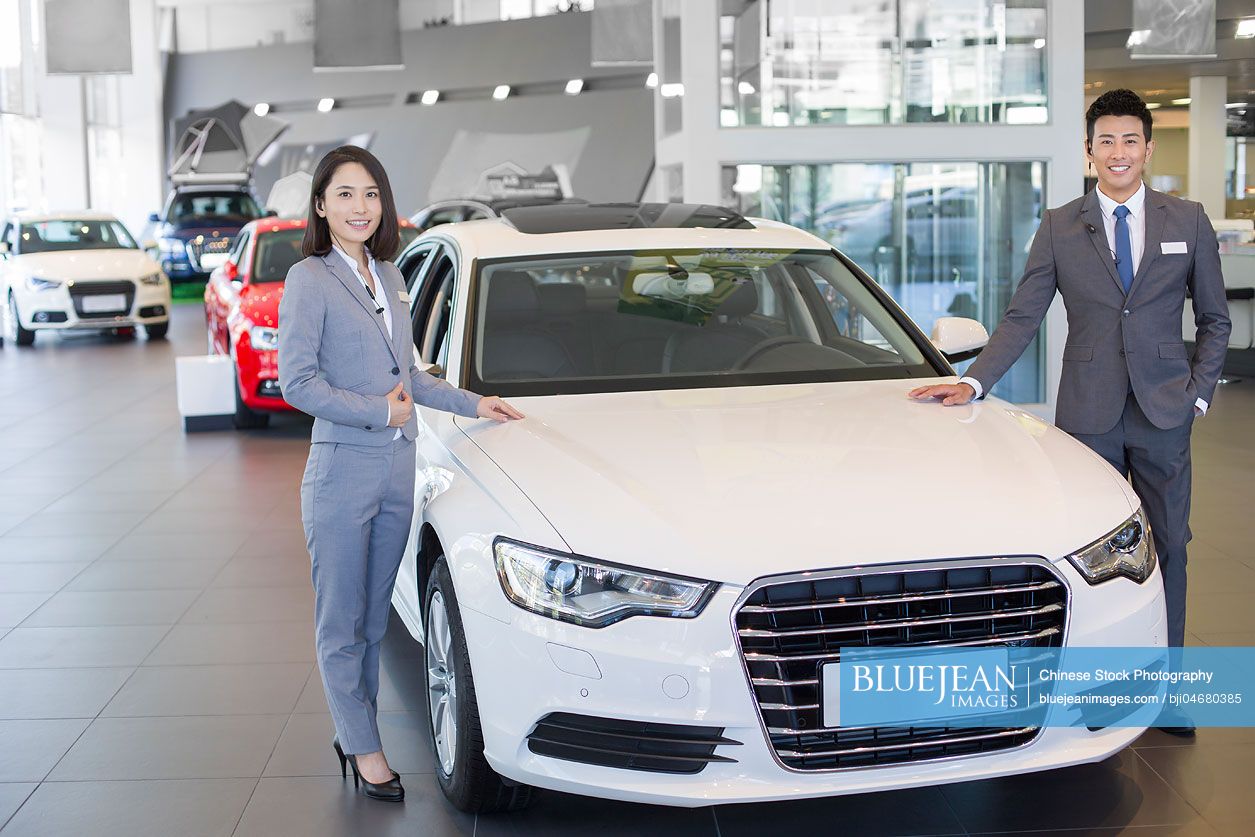 Confident Chinese salespeople standing with new cars in showroom