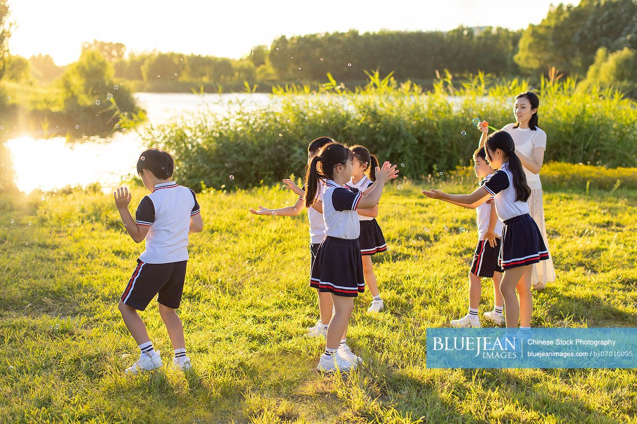 Cheerful Chinese school children and their teacher blowing bubbles on meadow
