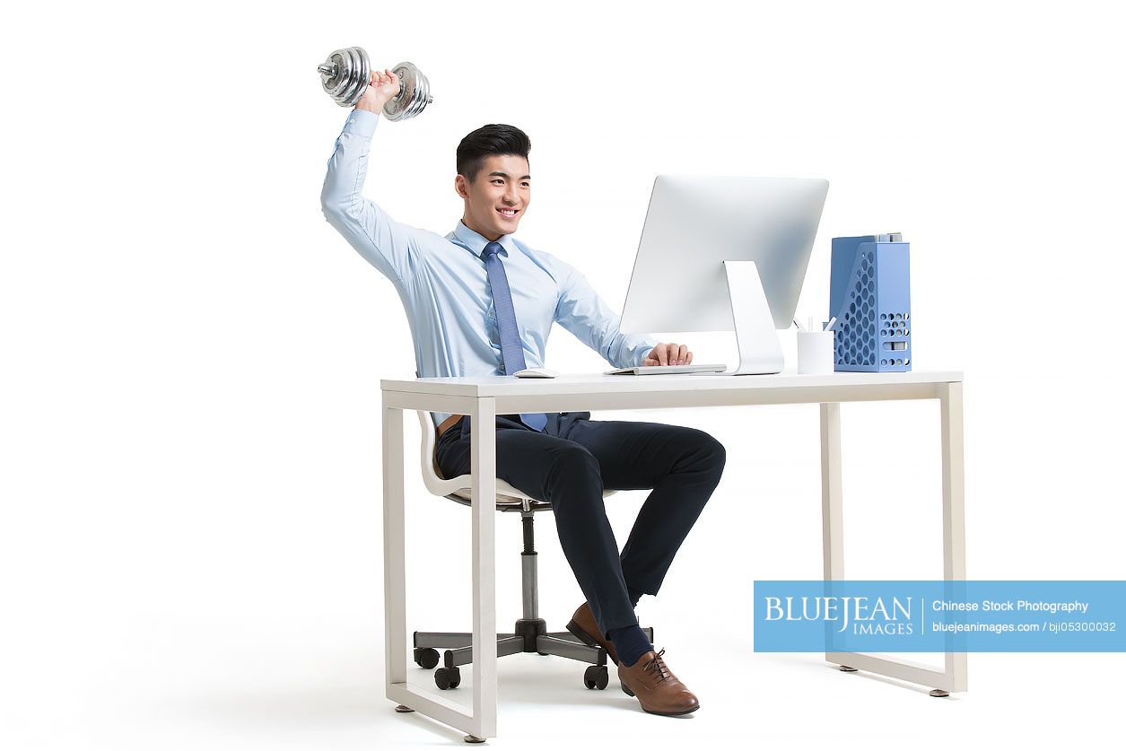 Young Chinese businessman lifting weights while using computer at work