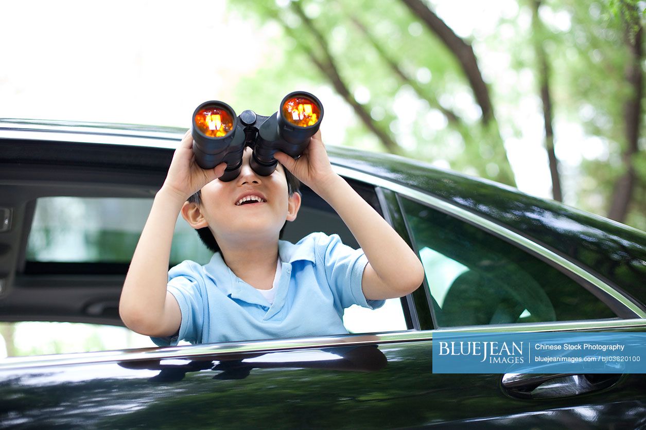 Cheerful Chinese boy leaning out of car window and looking with binoculars