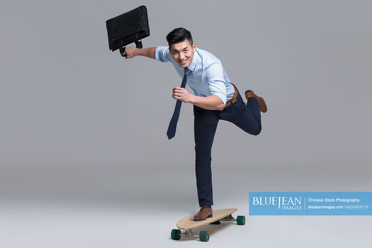 Cheerful young Chinese businessman skateboarding with briefcase