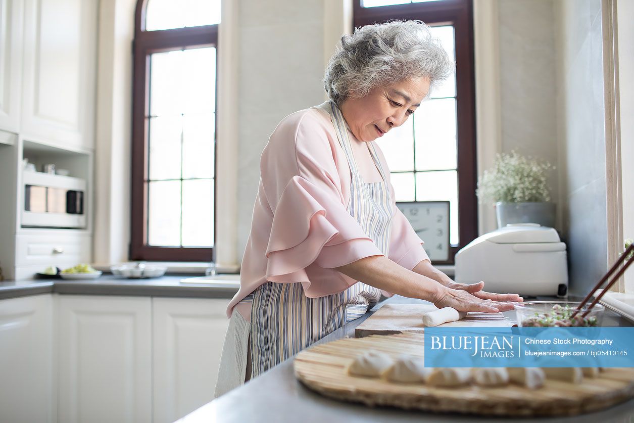 Cheerful senior Chinese woman making dumplings in kitchen