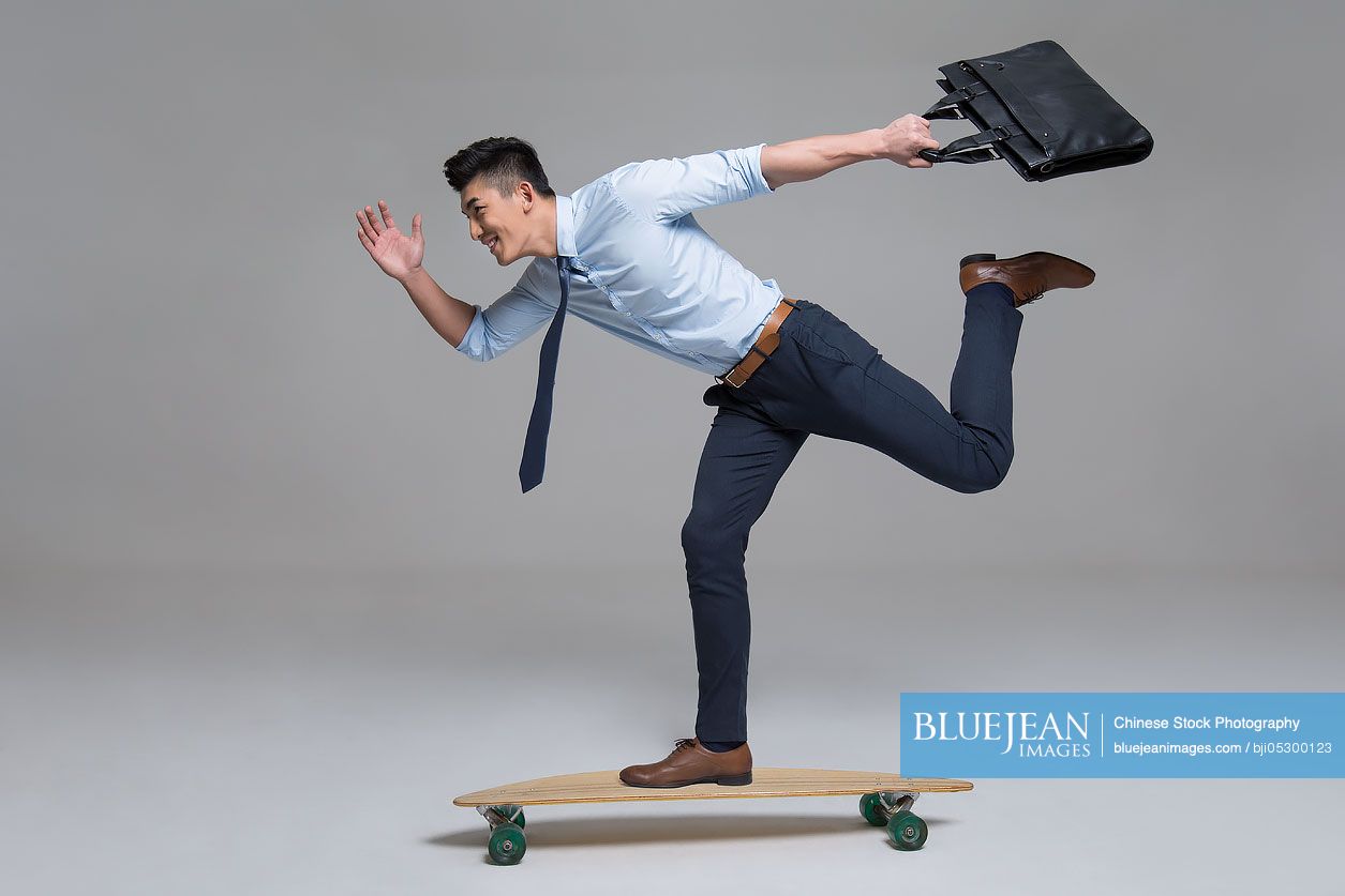 Cheerful young Chinese businessman skateboarding with briefcase