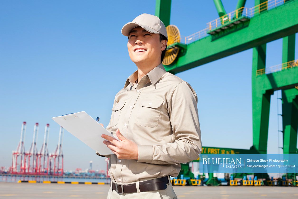 Male Chinese shipping industry worker with shipping dock and crane in the background