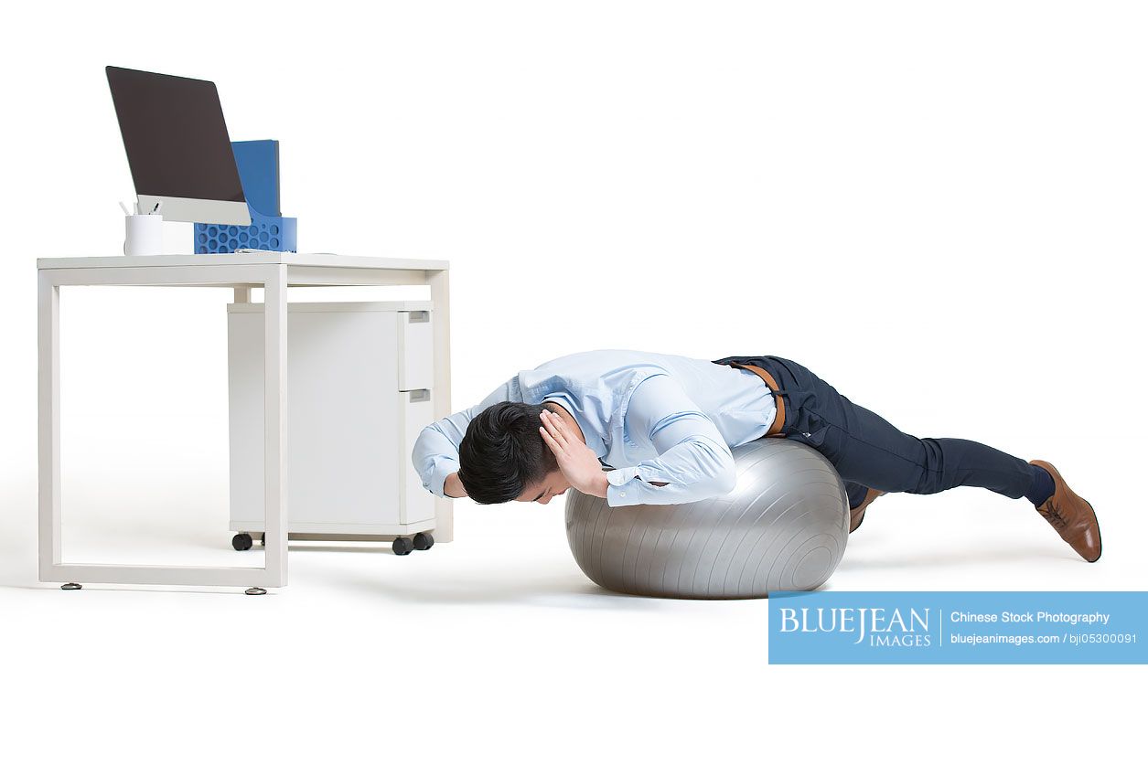 Young Chinese businessman exercising in office while sitting on fitness