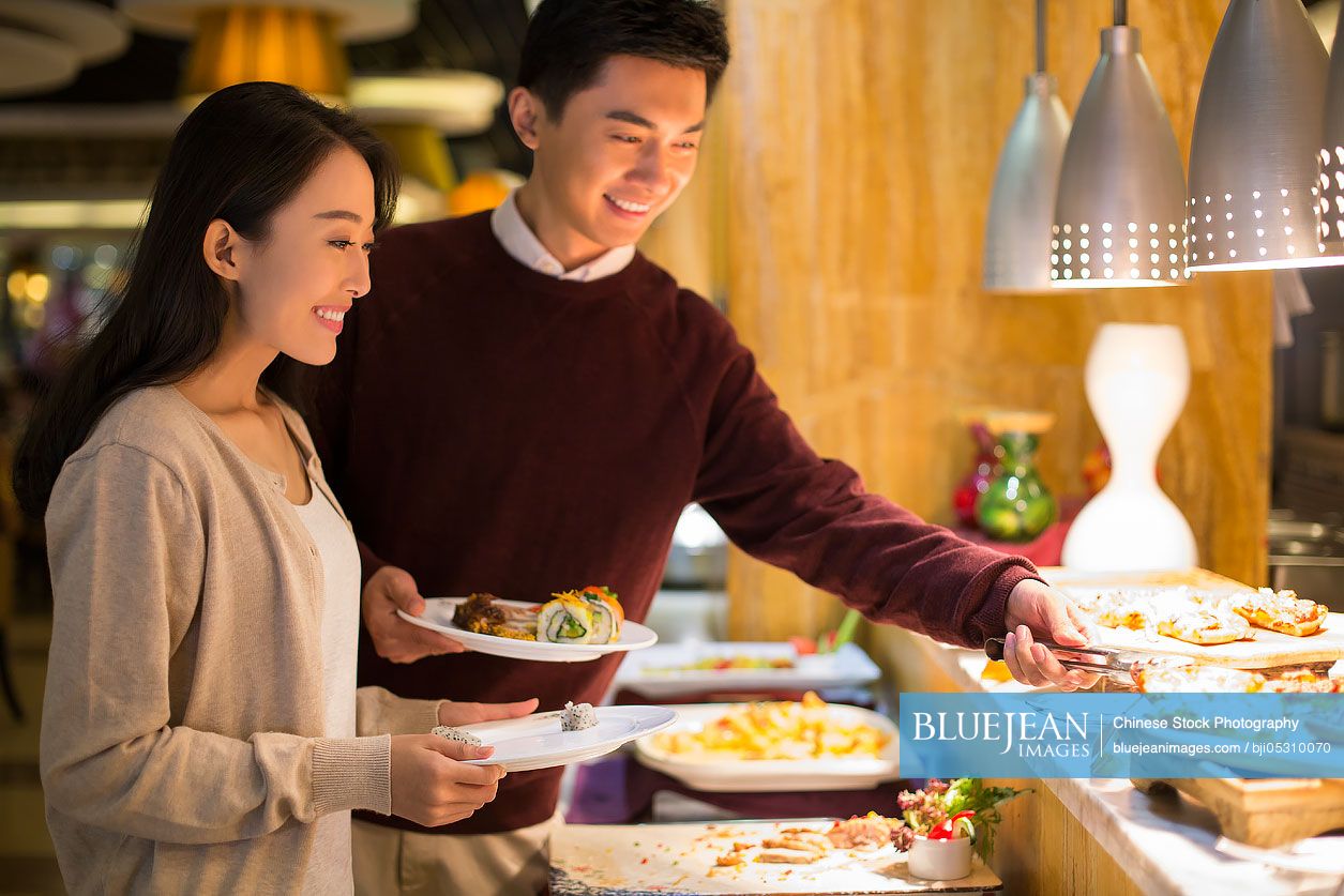 Cheerful young Chinese couple taking food from buffet table