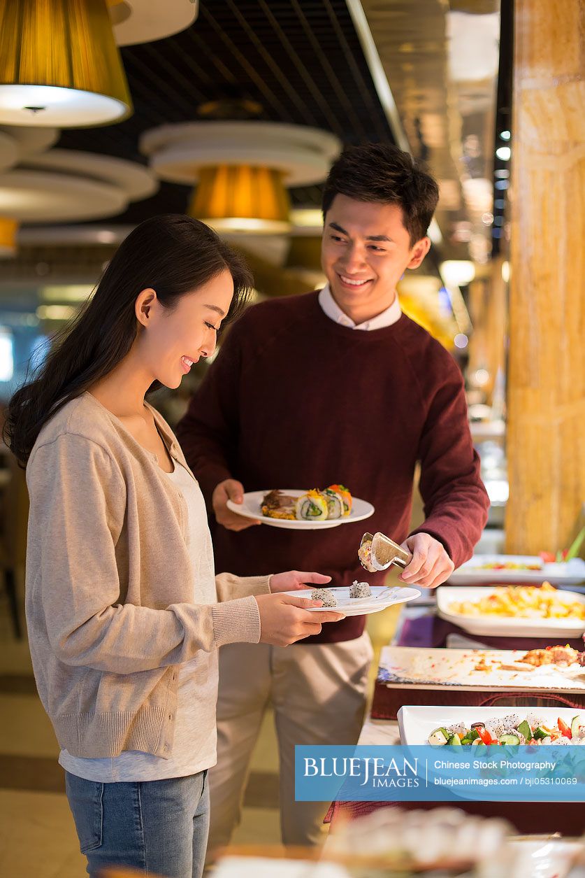 Cheerful young Chinese couple taking food from buffet table