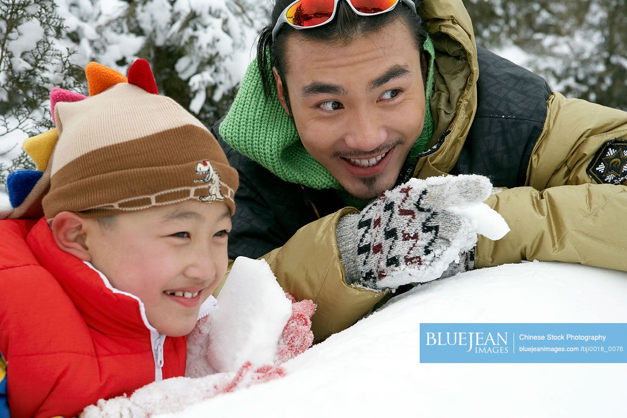 Chinese father and son making snowballs, hiding behind snow wall