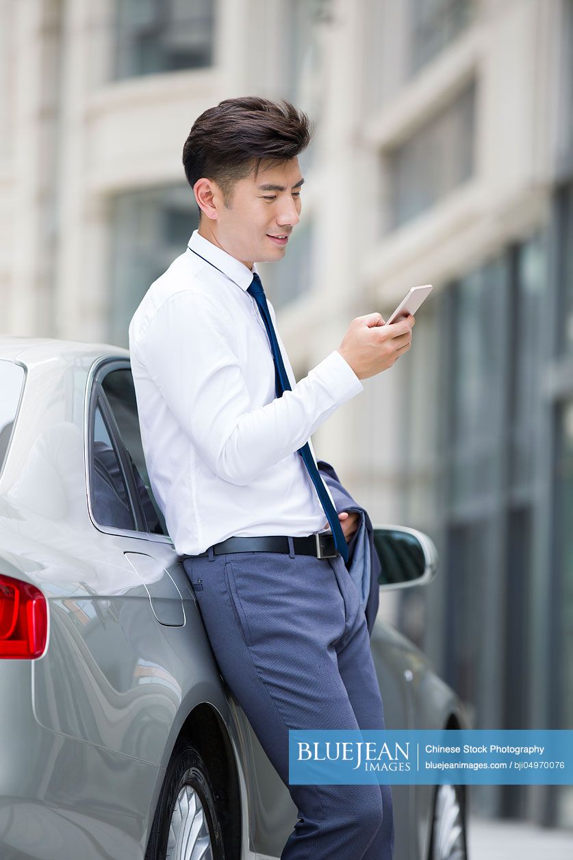 Confident Chinese businessman leaning on car with smart phone