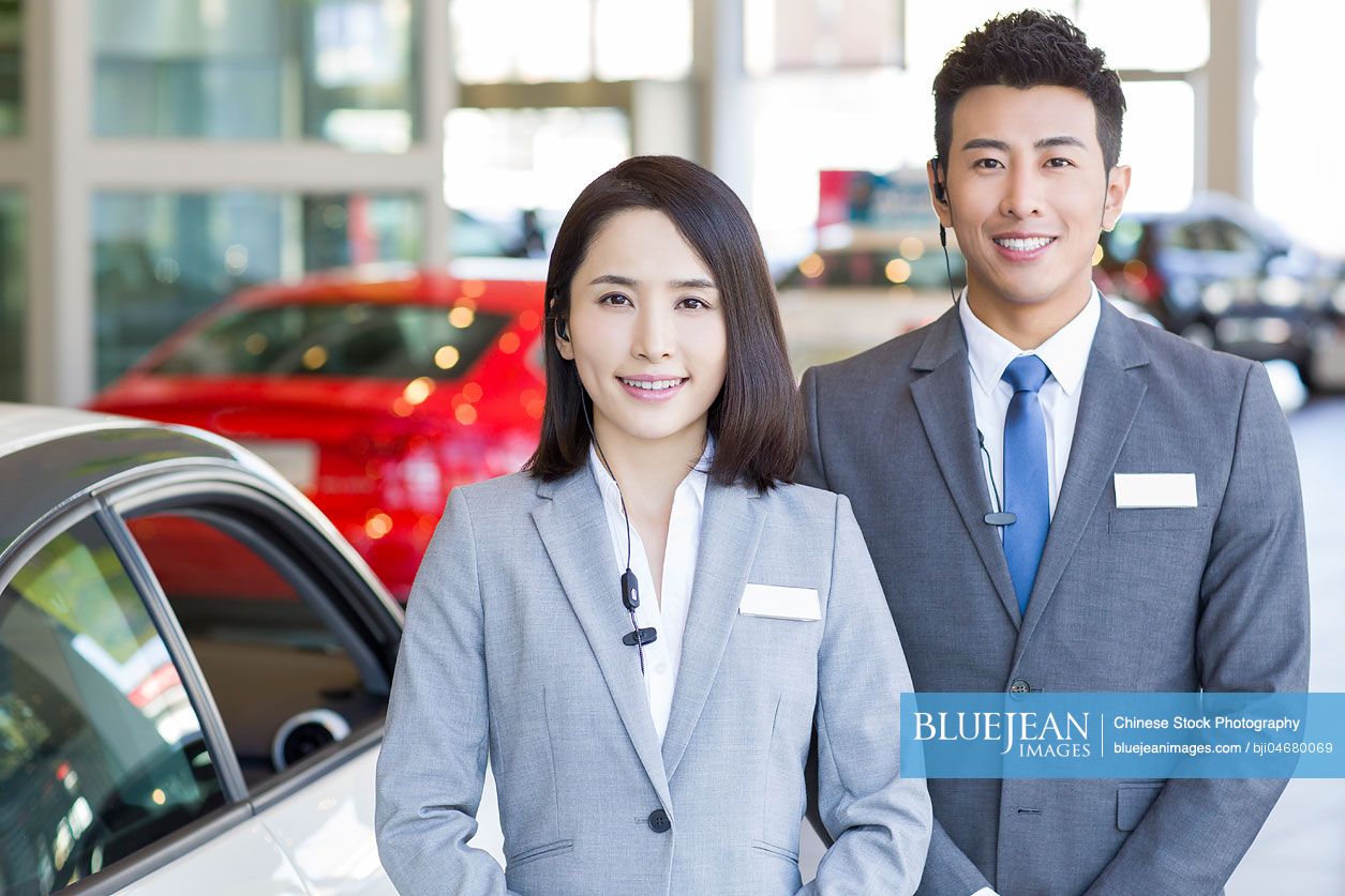 Confident Chinese salespeople standing with new cars in showroom