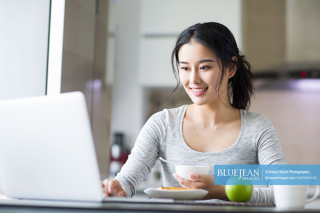 Young Chinese woman using laptop while having breakfast