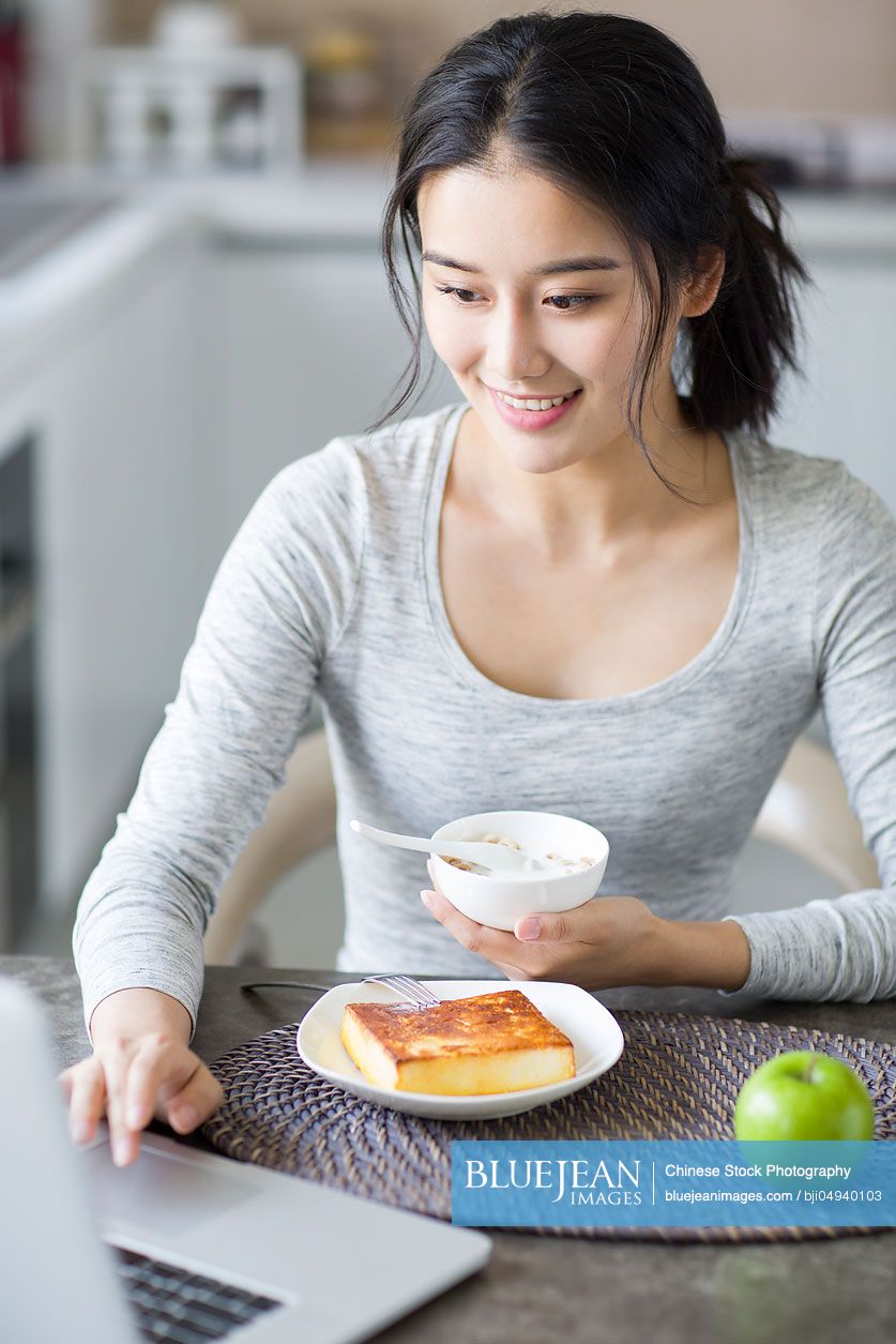 Young Chinese woman using laptop while having breakfast