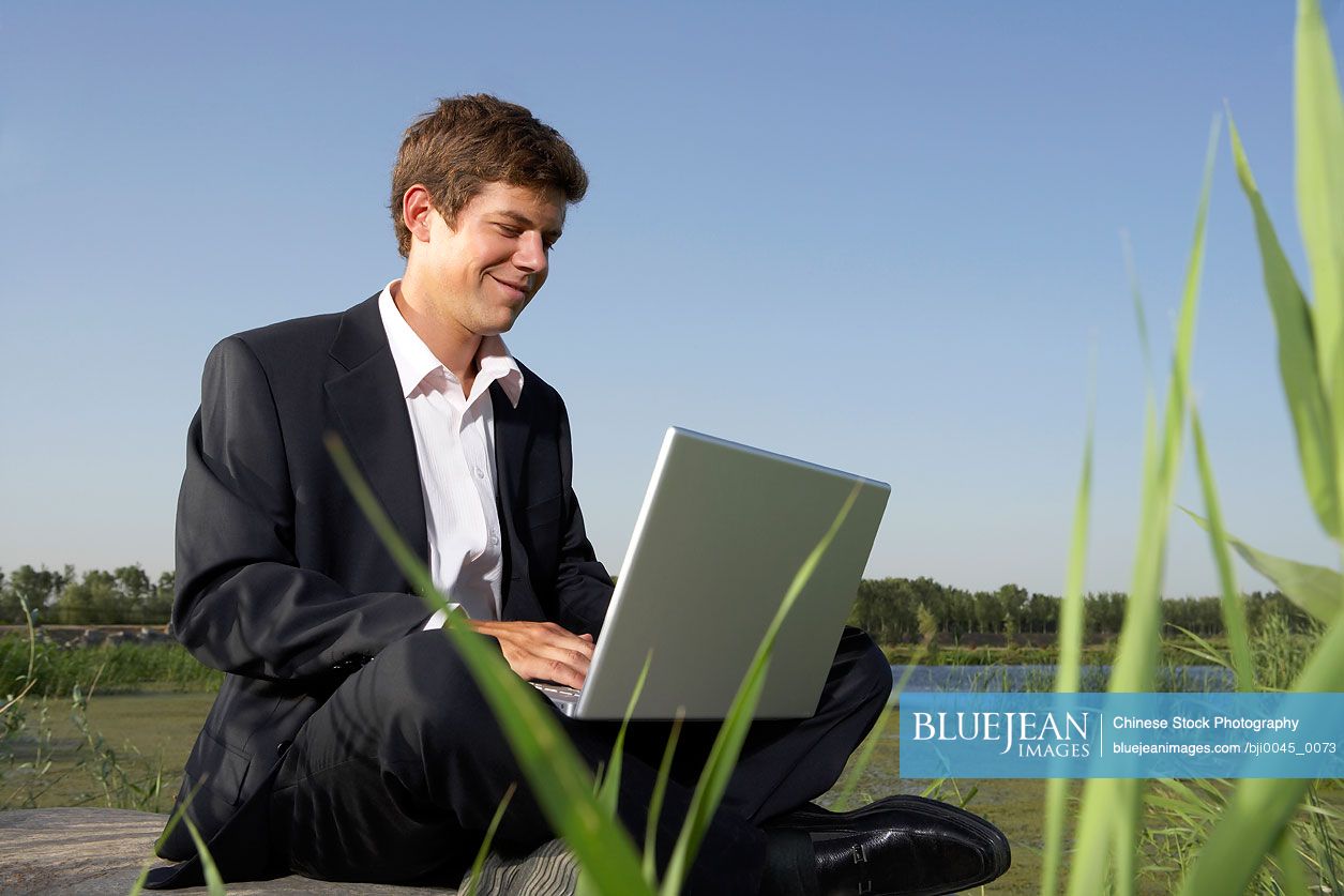 Chinese Businessman Sitting In A Field Using A Laptop Computer